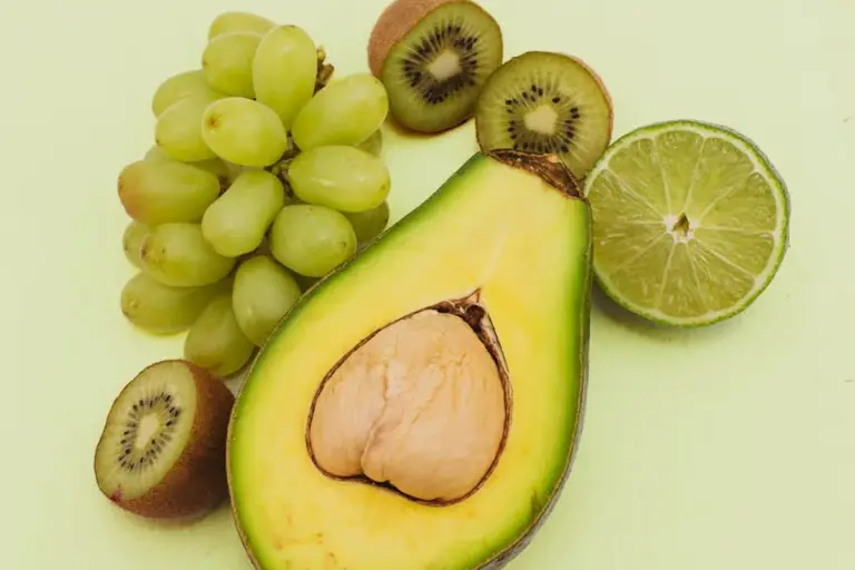 Halved avocado with seed visible, surrounded by grapes, kiwi slices, and a lime on a pale yellow background.