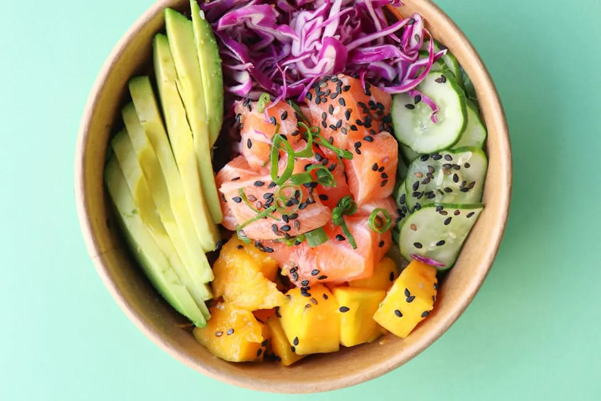 Colorful bowl of fresh foods arranged in sections: sliced avocado, mango chunks, cucumber, purple cabbage, salmon, and sesame seeds.