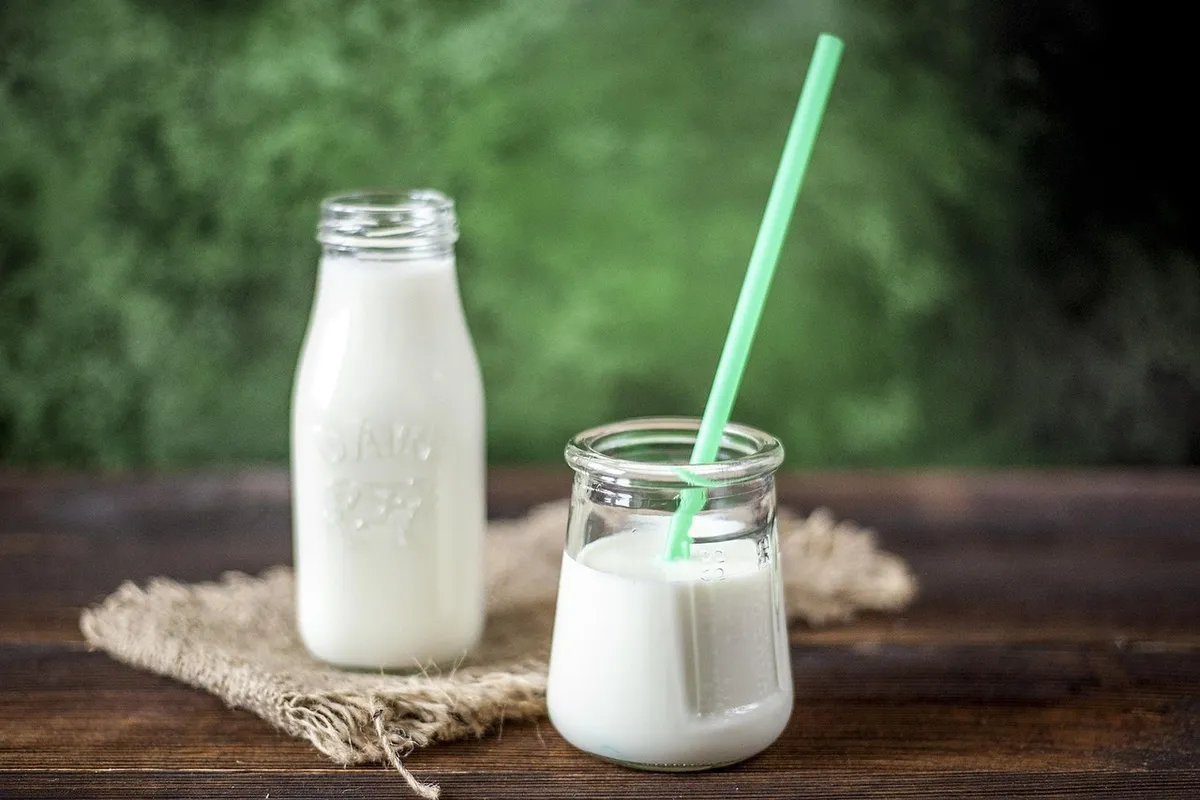 Two glass bottles of milk on a wooden surface with a green straw in the nearer bottle and a blurred green outdoor background.