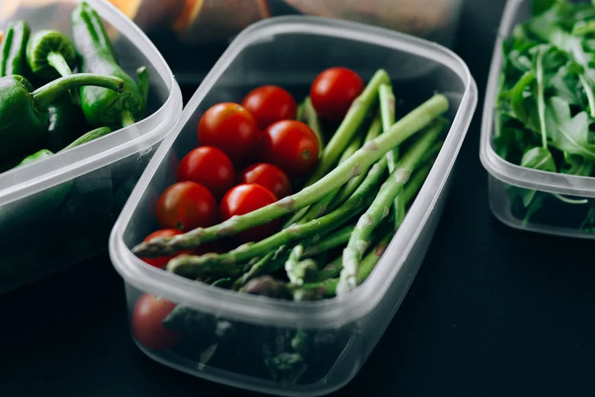 Clear plastic containers filled with fresh vegetables and greens (including asparagus, cherry tomatoes, and leafy greens) on a dark surface, ready to offer to a bearded dragon.