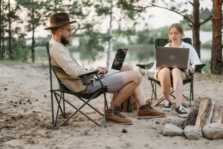 Two people sit outdoors in camping chairs with laptops, planning a bearded dragon's care before an owner's absence.
