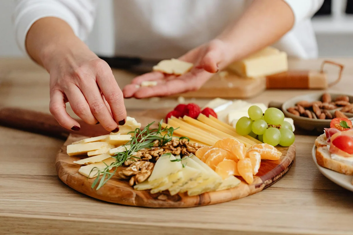 Hands arranging a wooden cheese board with cheeses, grapes, nuts, and herbs on a light wooden table.