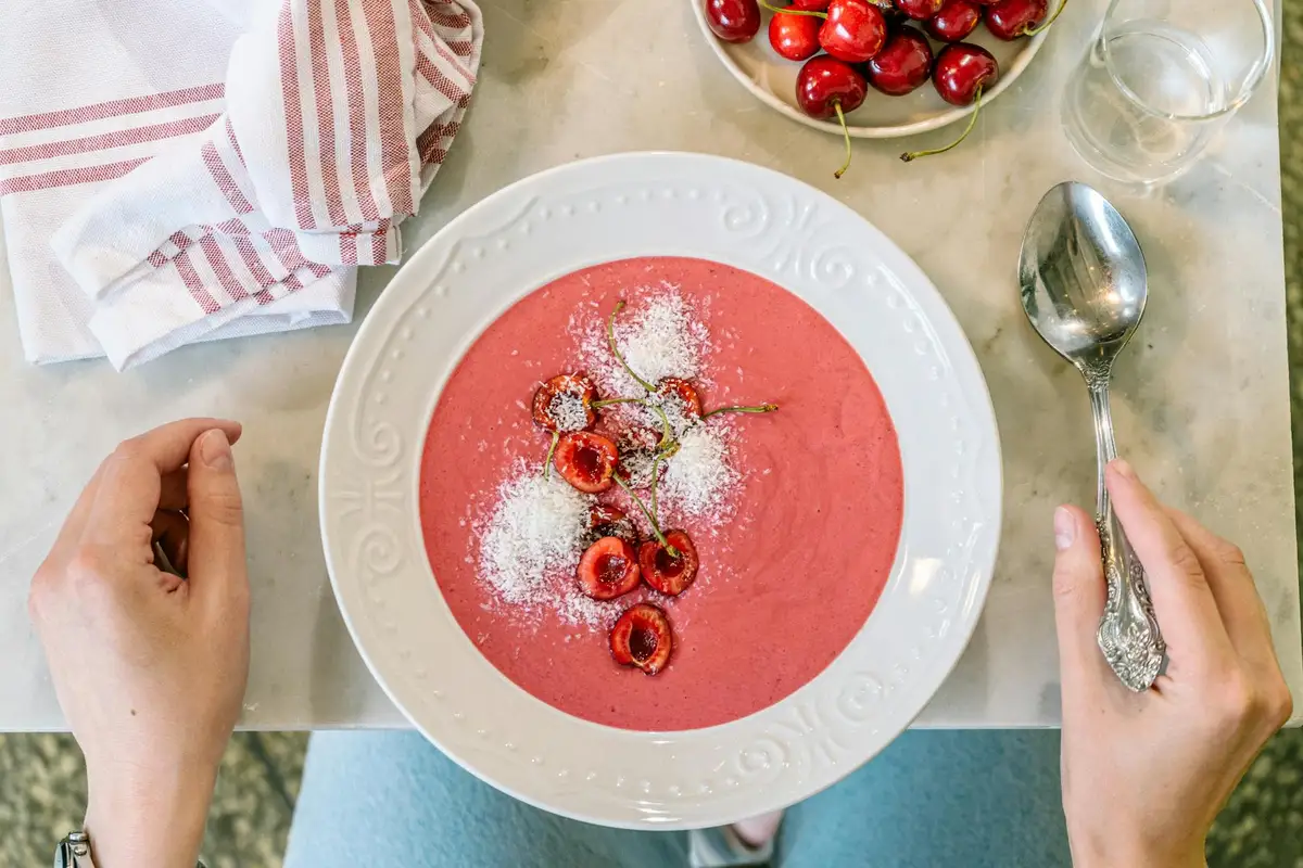 Top-down view of a pink cherry dessert bowl topped with halved cherries and powdered sugar, with a spoon to the right and hands near the plate.