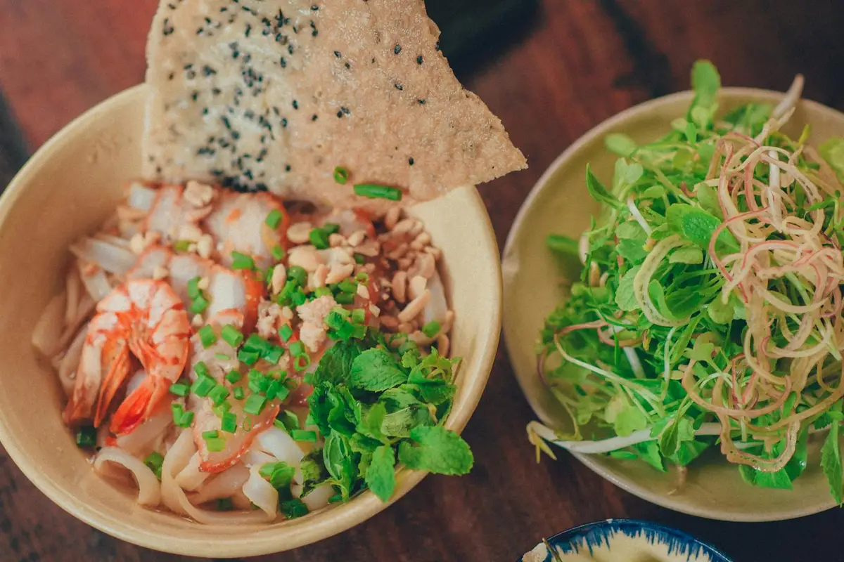 Two bowls of food on a wooden table: left bowl contains shrimp, noodles, herbs, and seaweed crisps; right bowl features a fresh green salad with herbs and shredded onions.