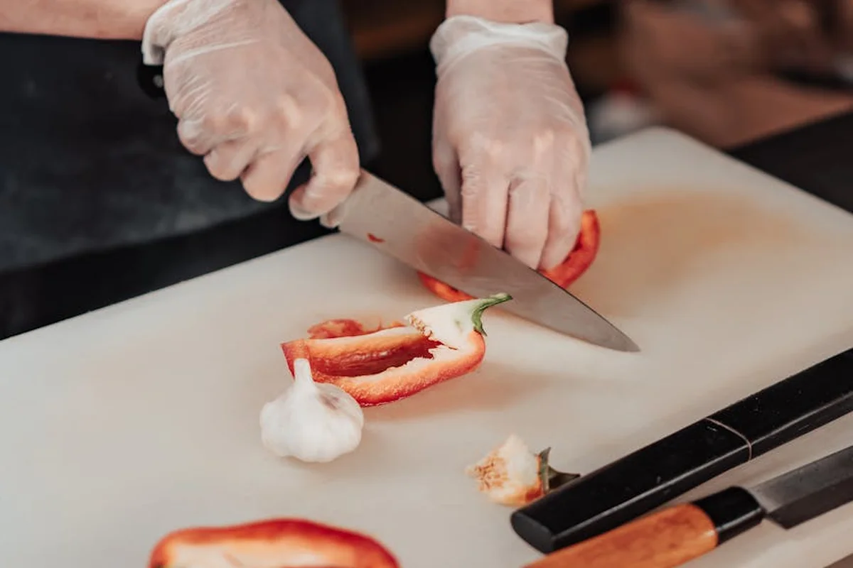 Close-up of gloved hands slicing a red bell pepper on a cutting board with a knife nearby.