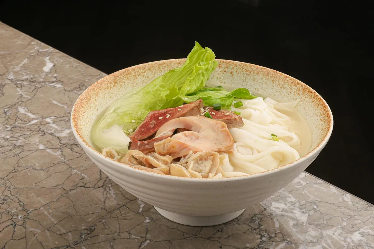 Bowl of noodle soup with leafy greens and sliced meat, showing fresh greens similar to bok choy.