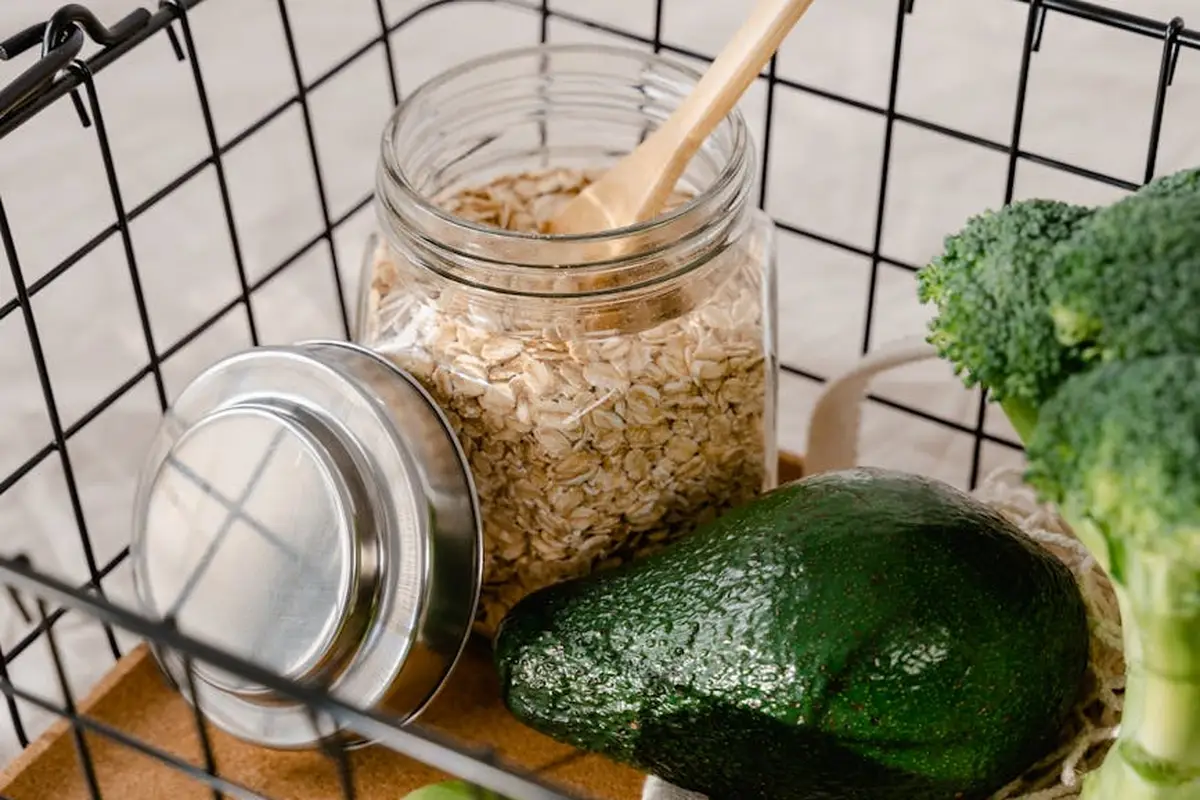 Jar of oats with a wooden spoon, a head of broccoli, and a dark green avocado on a wooden surface inside a metal grid backdrop