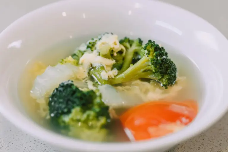 A white bowl containing broccoli florets and stalks in a light broth with a small carrot piece, photographed close-up.