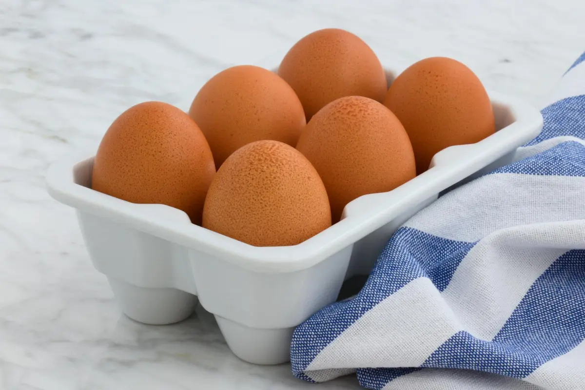 Six brown eggs in a white egg carton with a blue striped cloth beside them.