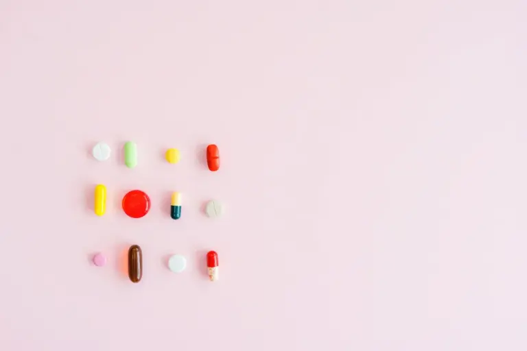 Assorted colorful pills and capsules arranged on a light pink background.