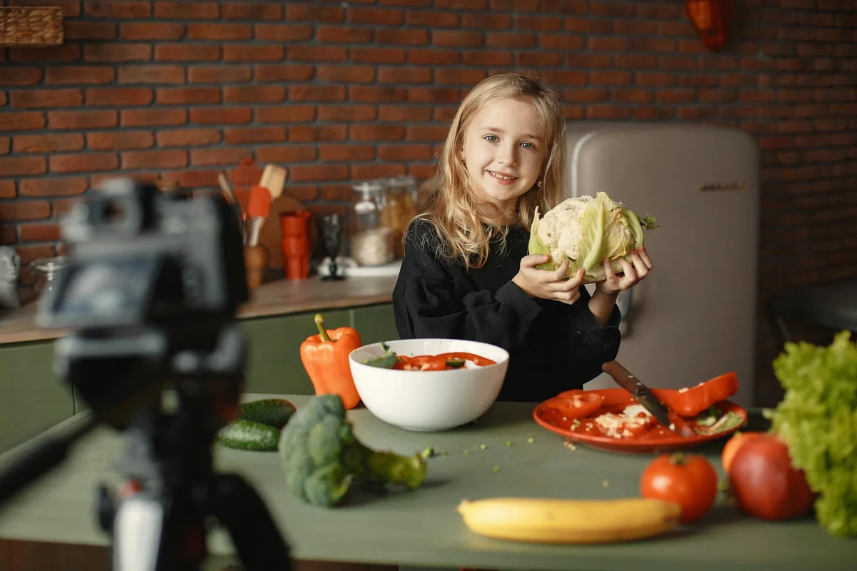 A young girl in a kitchen holds a head of cauliflower, with a bowl of salad and vegetables on the counter and a camera filming in the foreground.