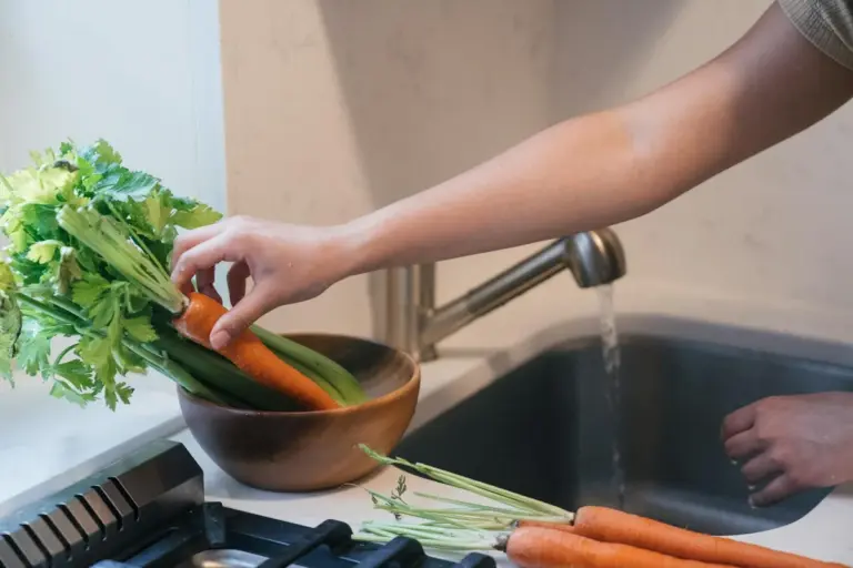 Person rinsing and preparing celery stalks and greens on a kitchen counter with carrots nearby