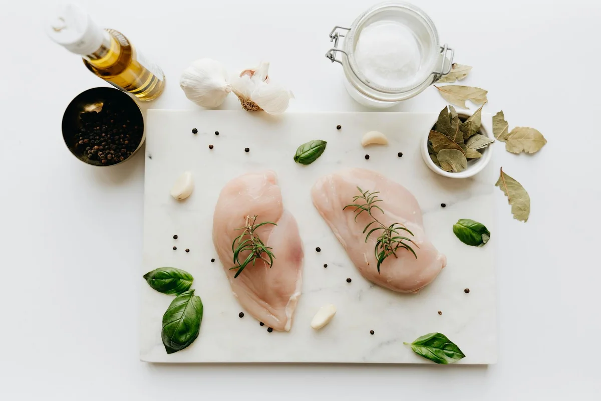 Two raw chicken breast fillets on a white cutting board with garlic, herbs, and peppercorns.