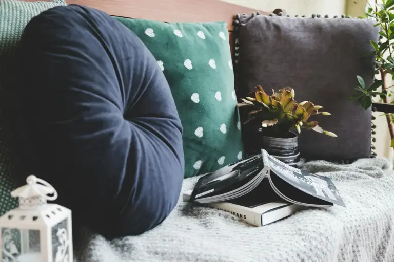 Cozy reading corner with a navy circular pillow, a green polka-dot pillow, a succulent plant, an open magazine, and a white lantern on a soft knit blanket.