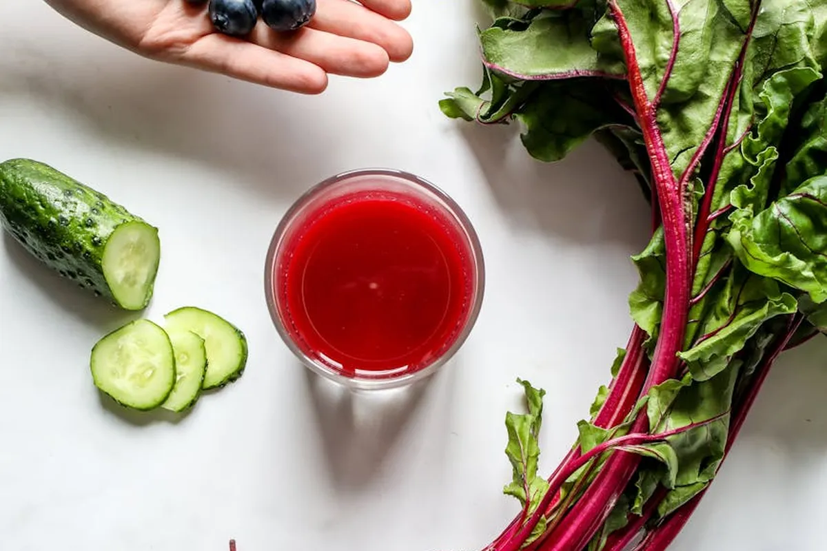 Cucumber slices, a glass of red juice, and fresh beet greens with purple-red stems on a white surface.