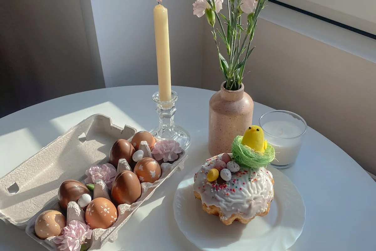 A kitchen table showing a carton of brown eggs, a decorated cake with a yellow chick on top, a tall candle in a holder, and a vase with flowers.