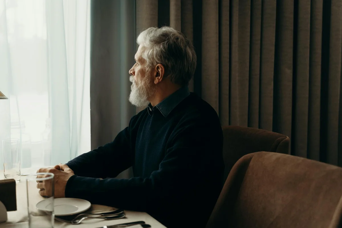Older man with a white beard sitting at a window-side table, looking outside.