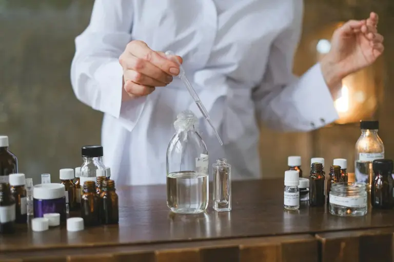 A person in a white lab coat uses a pipette to handle a liquid in a glass jar, surrounded by small vials and bottles on a wooden table.