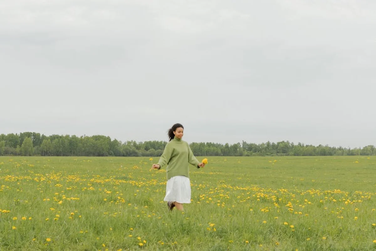 A woman in a light green sweater and white skirt walks through a field dotted with yellow dandelions under a cloudy sky.