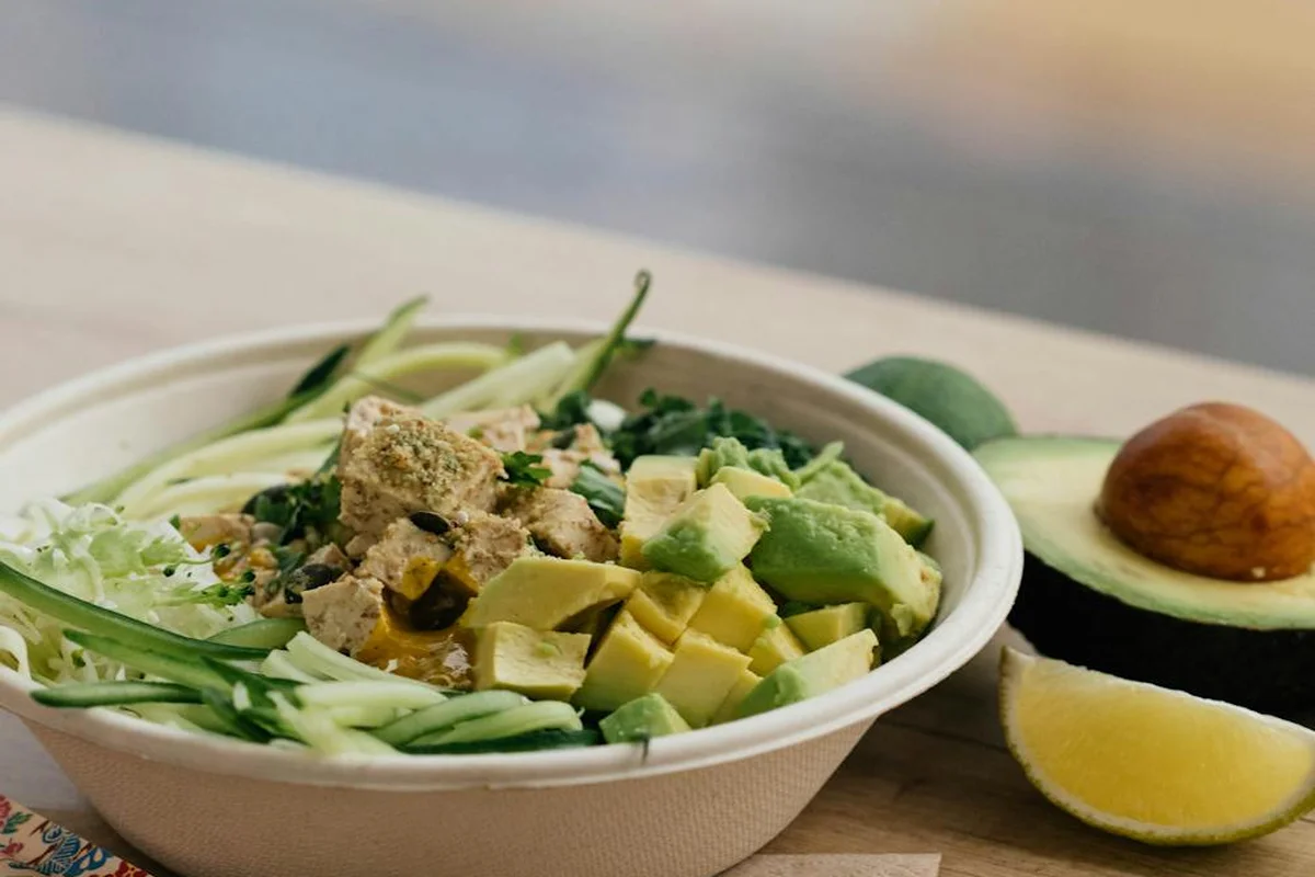 A bowl of chopped greens with cucumber ribbons and avocado chunks on a wooden surface, with avocado halves in the background.