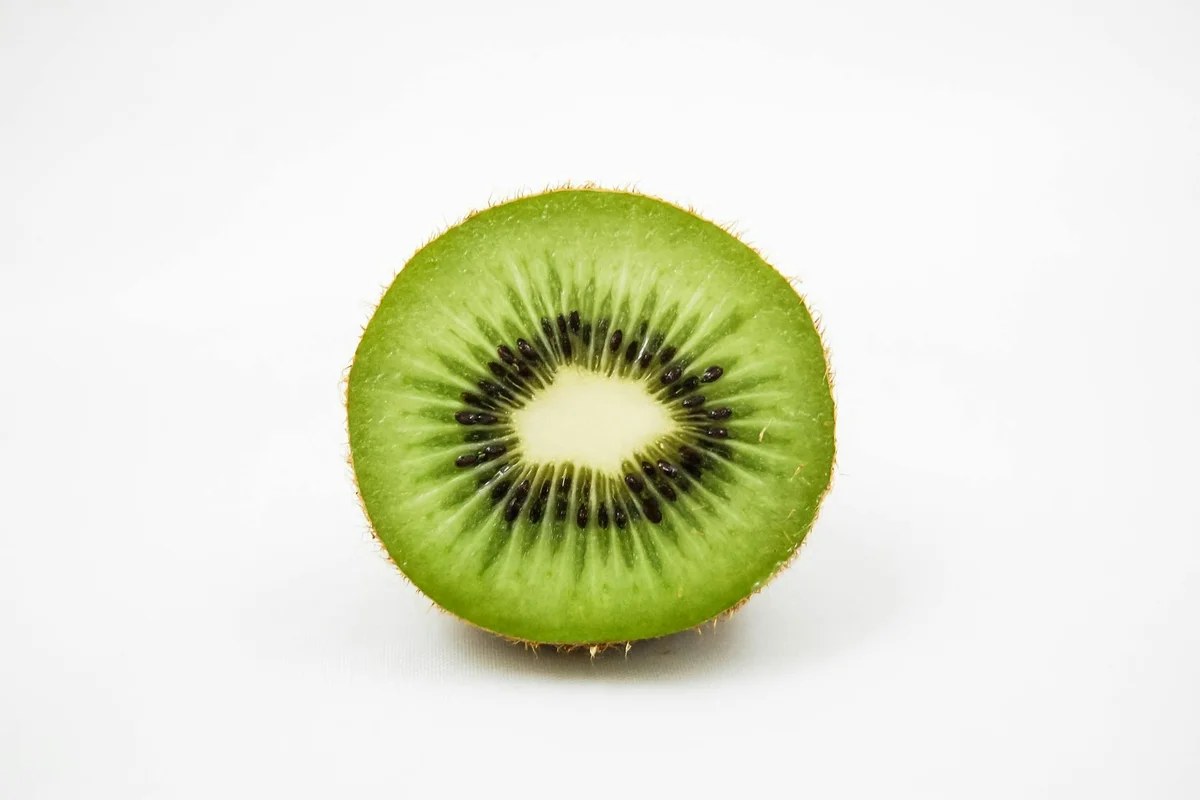 Cross-section of a green kiwifruit showing vibrant green flesh, small black seeds arranged in a ring around a pale center, with a fuzzy brown skin along the edge.