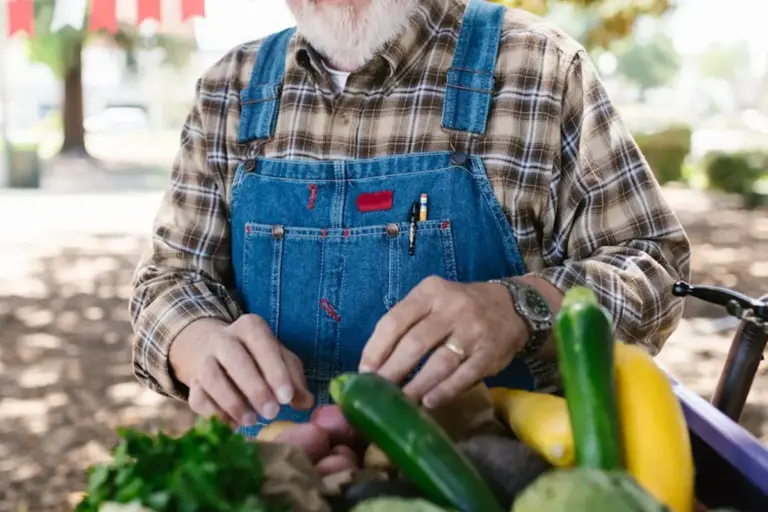 Older man with a gray beard wearing a plaid shirt and blue denim overalls preparing fresh vegetables at an outdoor market.