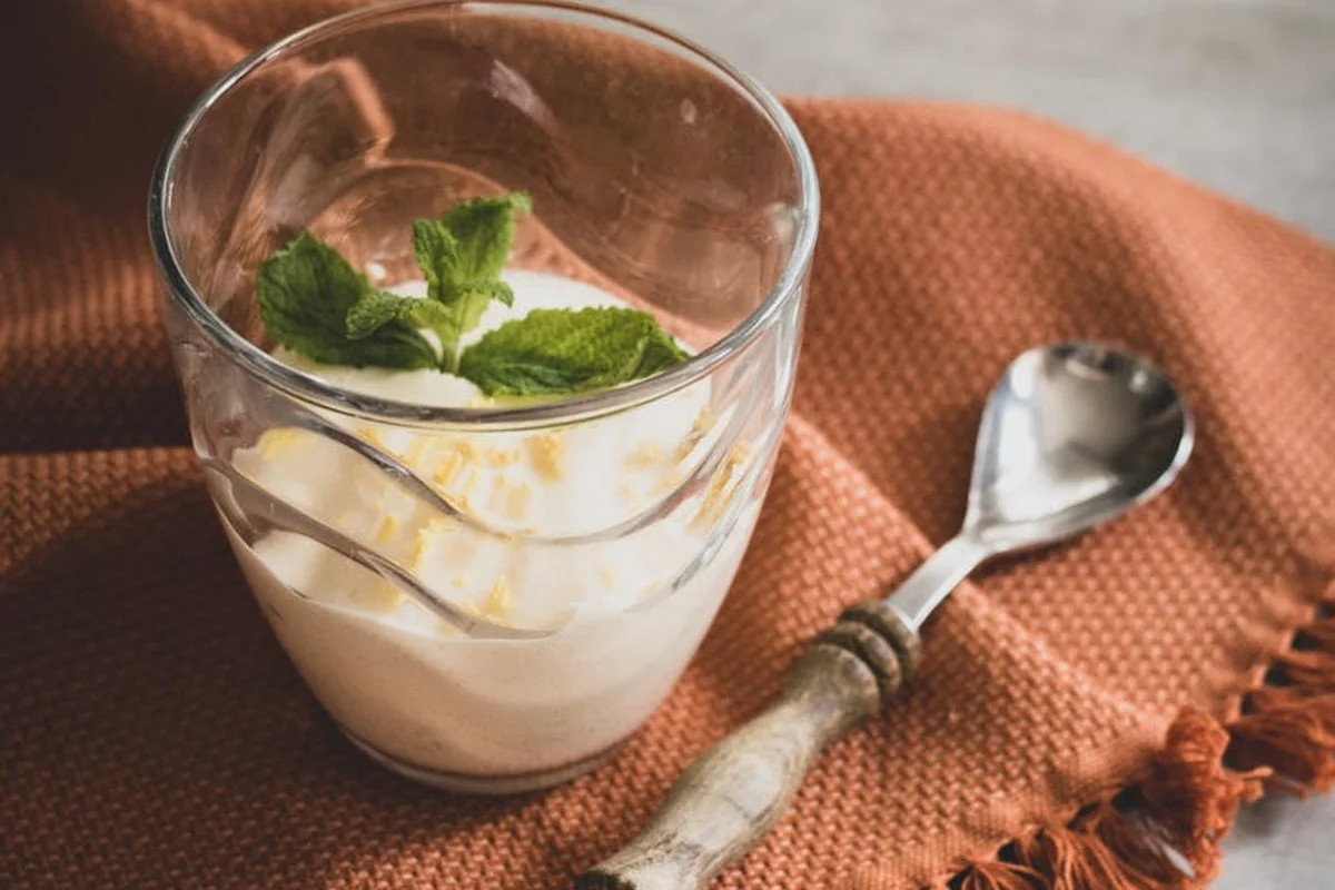 Glass of yogurt topped with fresh mint leaves on a textured orange cloth, with a spoon nearby.