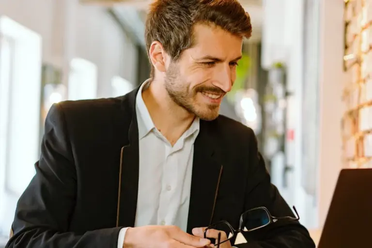 Man in a suit sits at an office desk, smiling, with eyeglasses in hand and a laptop in front.