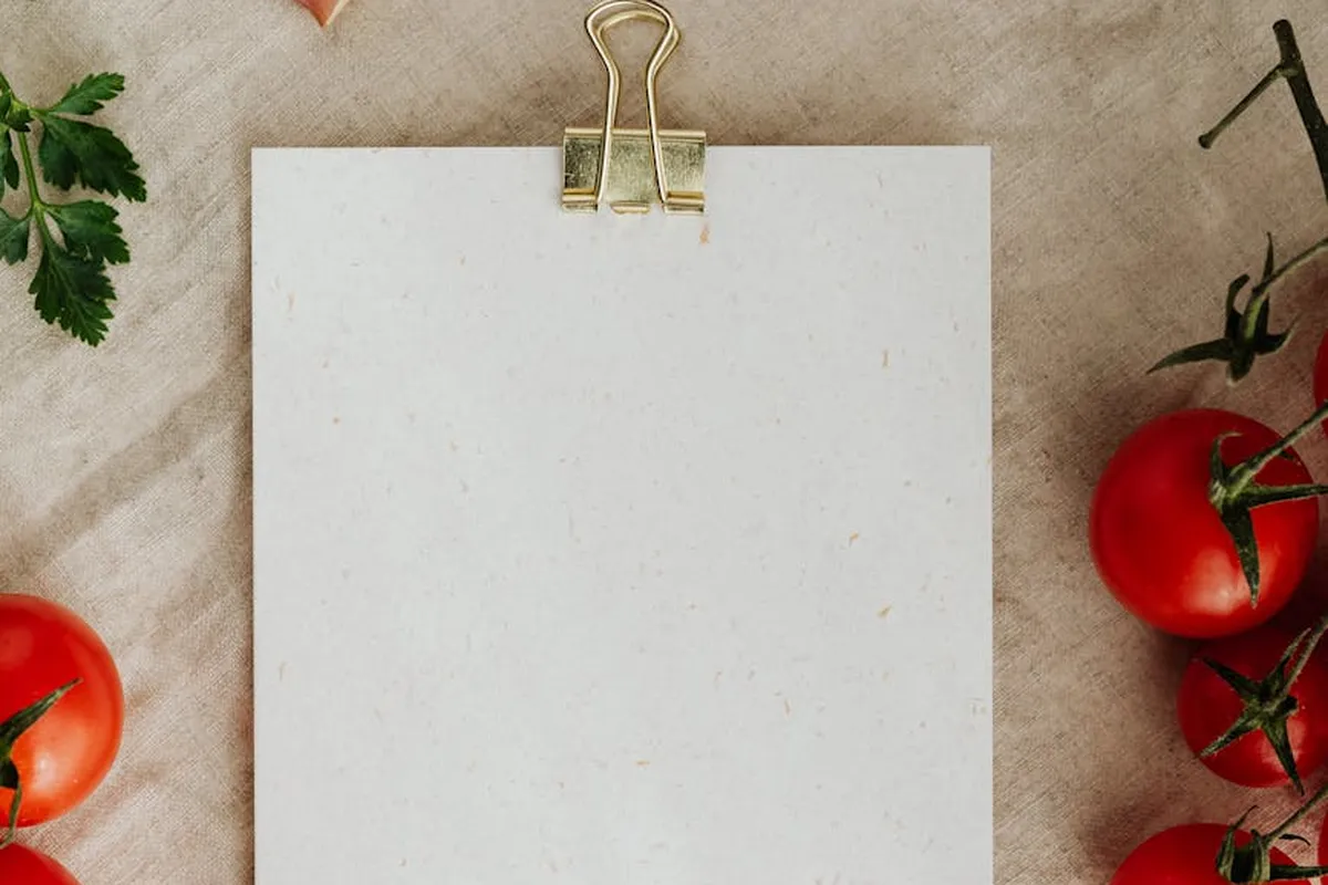 A blank white card clipped with a gold binder on a beige surface, with parsley sprigs in the corner and ripe tomatoes framing the scene.