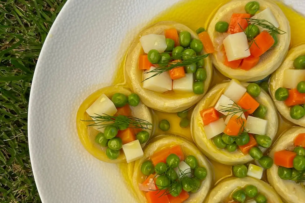 Close-up of round bite-sized appetizers topped with green peas, diced orange carrots, and white cheese cubes on a white plate, garnished with dill.