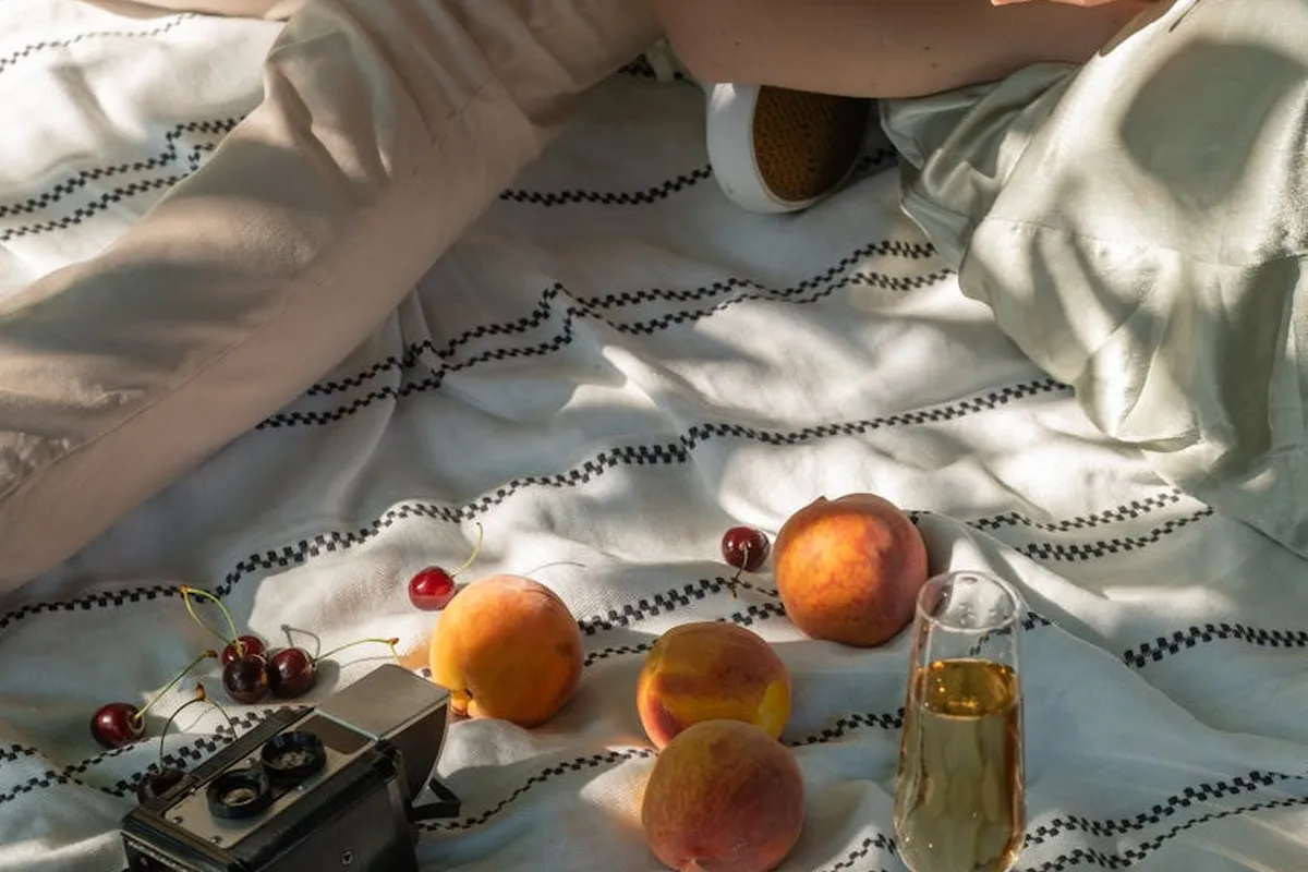 Peaches and other stone fruits on a striped blanket with a glass of juice and a camera nearby, setting a still-life scene for an article about alternative fruits for a balanced bearded dragon diet.