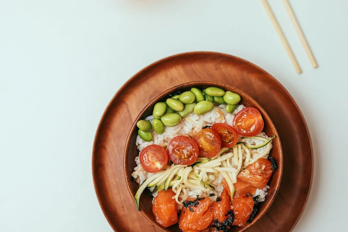 Wooden bowl on a pale blue background filled with fresh vegetables, including green edamame beans, sliced cherry tomatoes, and shredded zucchini, with chopsticks resting nearby.