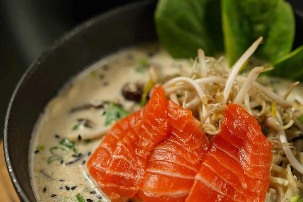 Salmon slices and a mound of bean sprouts in a creamy sauce served in a dark bowl, with greens in the background.
