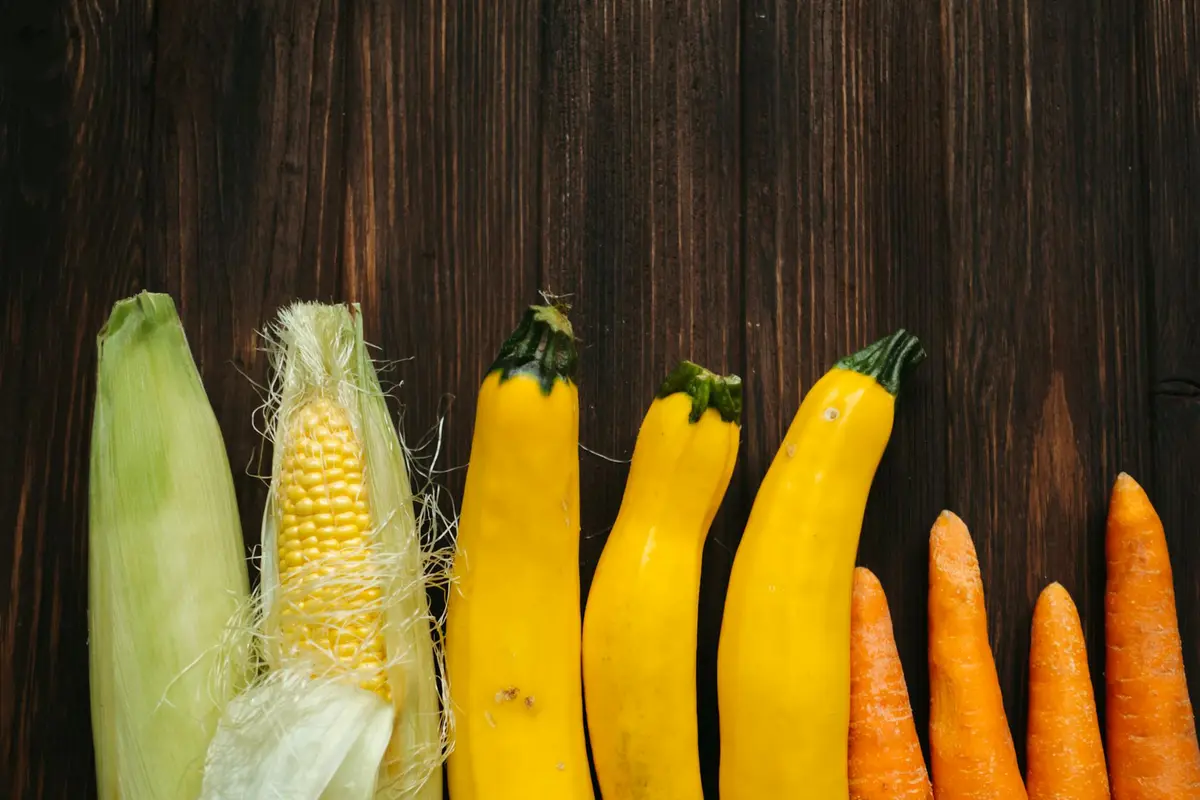 Row of yellow squash varieties on a dark wooden surface, with corn and carrots visible in the background.