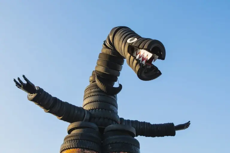 Dragon sculpture made from stacked tires reaching its neck upward against a clear blue sky.