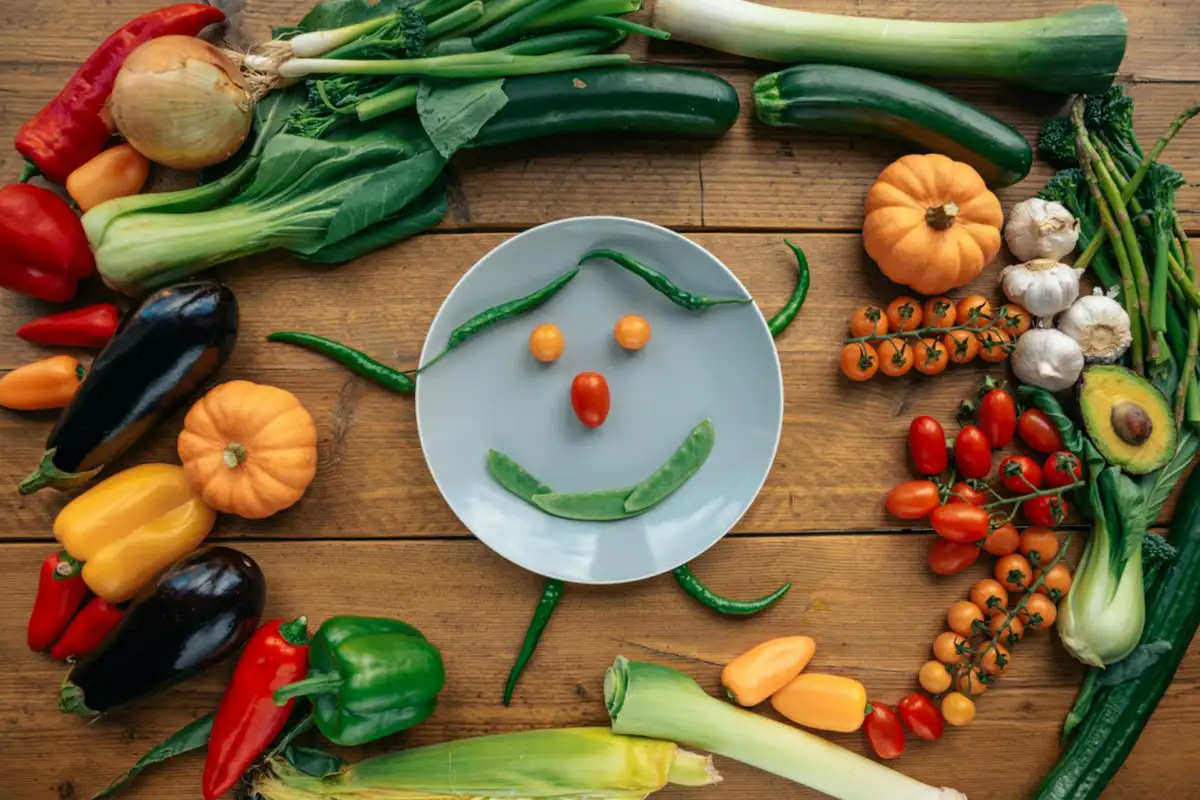 Assorted vegetables and greens arranged around a white plate on a wooden table, with a smiley face made from peppers in the center.