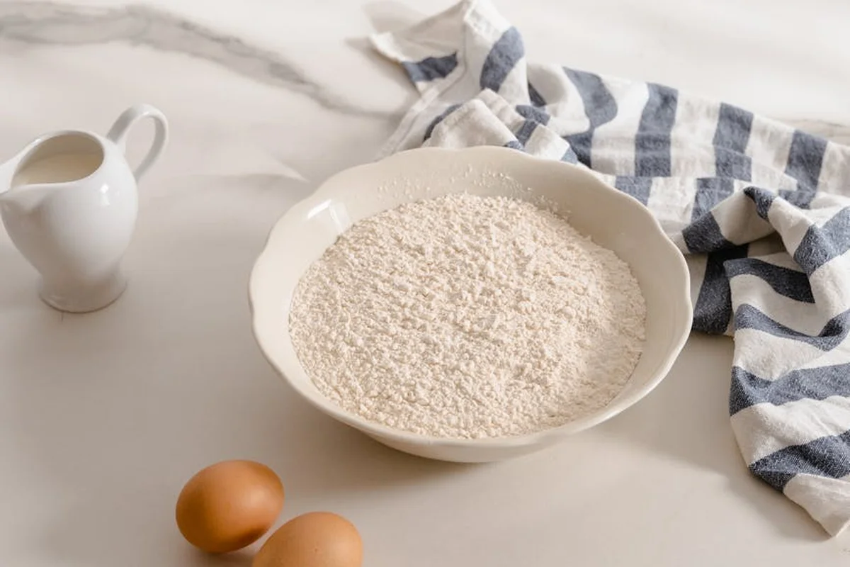 A white bowl filled with flour sits on a light countertop, with two eggs nearby and a small pitcher of milk, a striped cloth draped in the background.