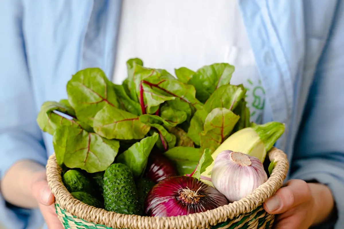A person holding a wicker basket filled with fresh vegetables, including red and white onions, leafy greens, garlic, and cucumbers.