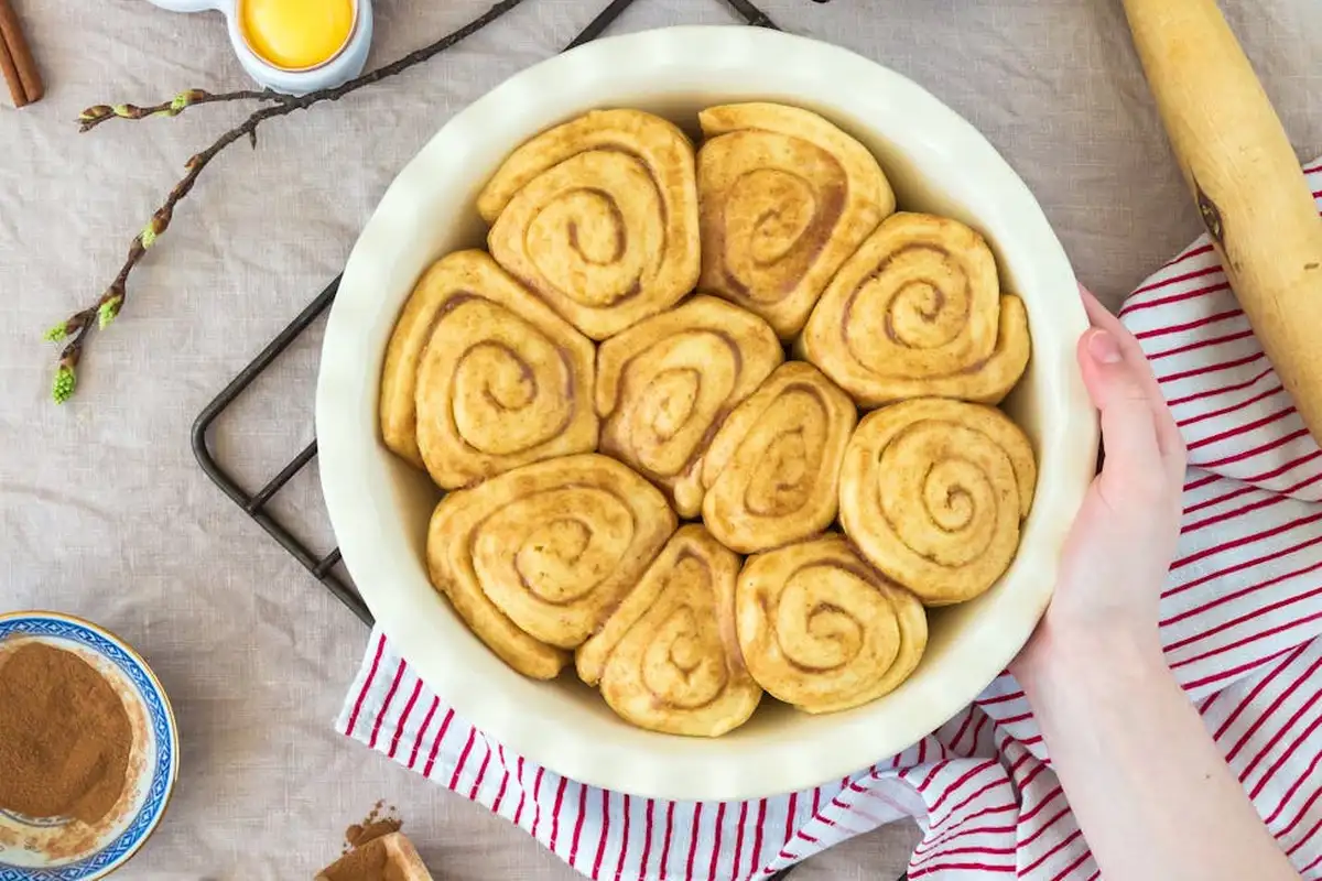 A white round plate filled with cinnamon rolls on a light-colored table, with a hand resting on the plate and a striped napkin nearby.
